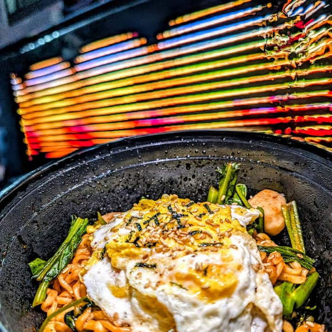 Noodles with greens and a fried egg on top, in a black bowl on the griddle. Photograph posted by Hasina's Burmese Kitchen.